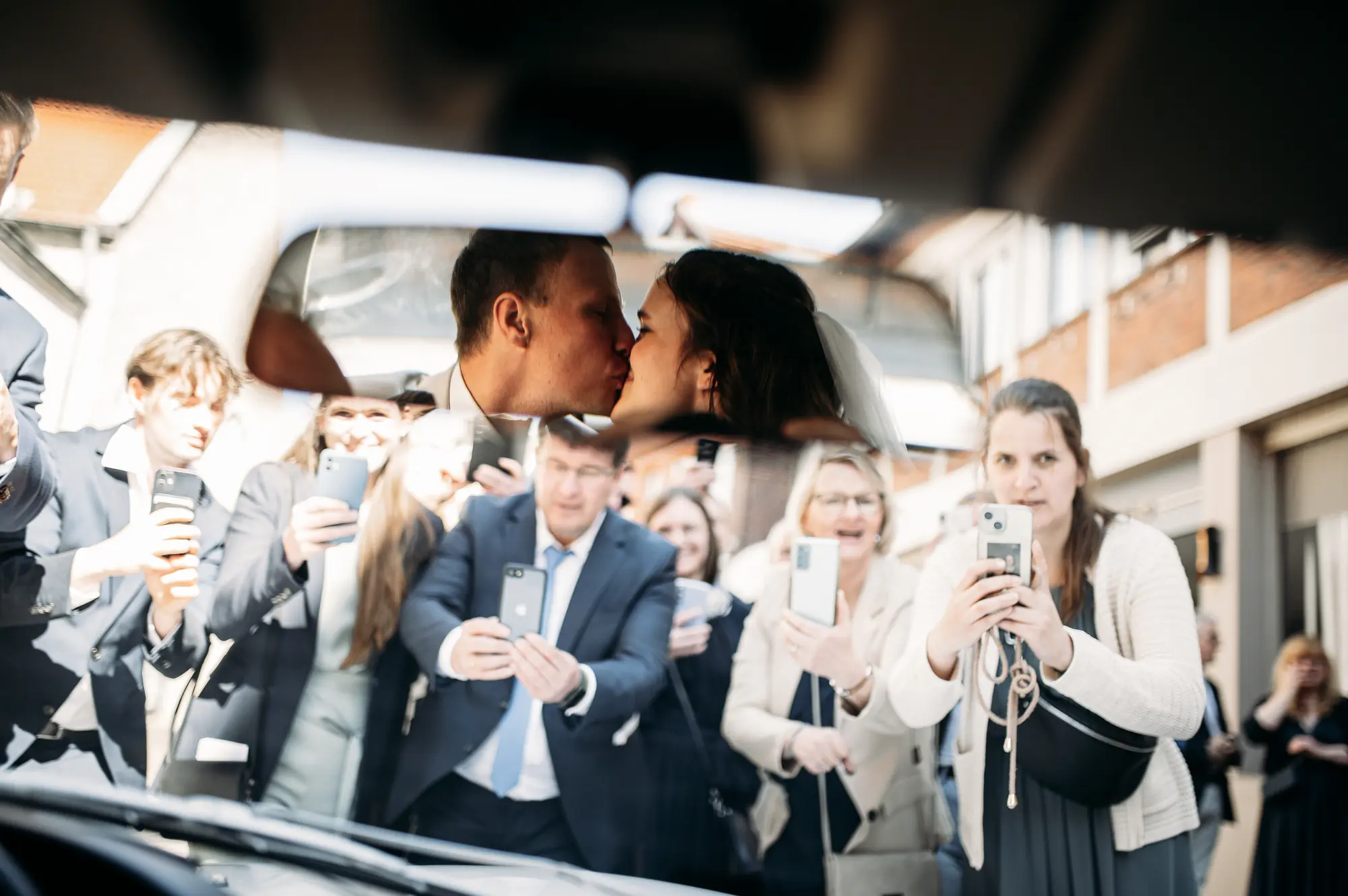 Kuss im Rückspiegel des Hochzeitsautos, während Gäste den Moment mit dem Smartphone festhalten