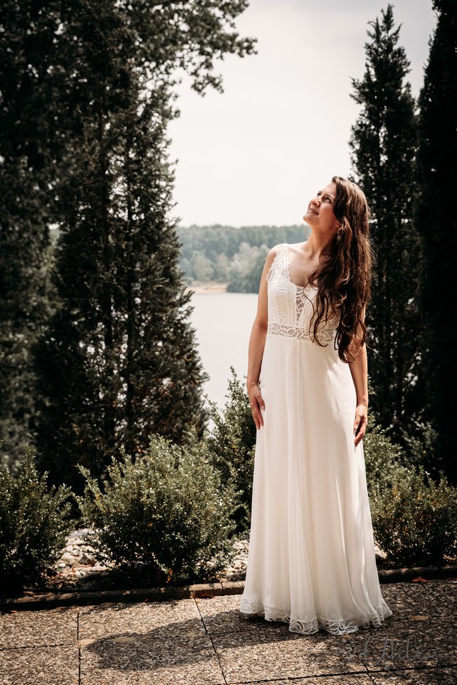 Braut in weißem Kleid blickt lächelnd nach oben zwischen Zypressen an einem See