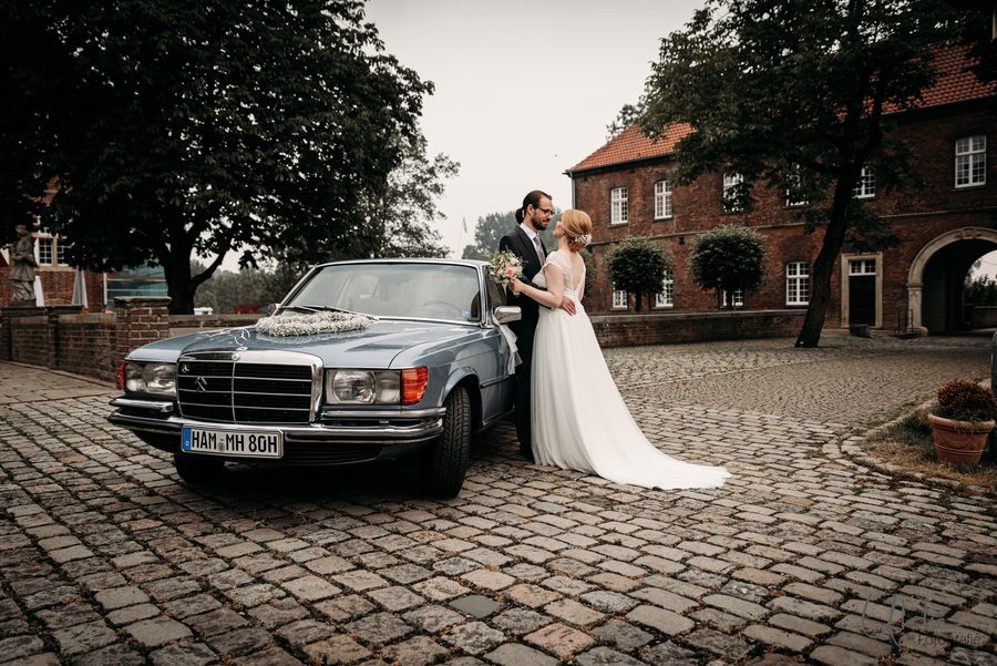 Brautpaar steht neben einem Oldtimer-Hochzeitsauto vor einem Backsteingebäude